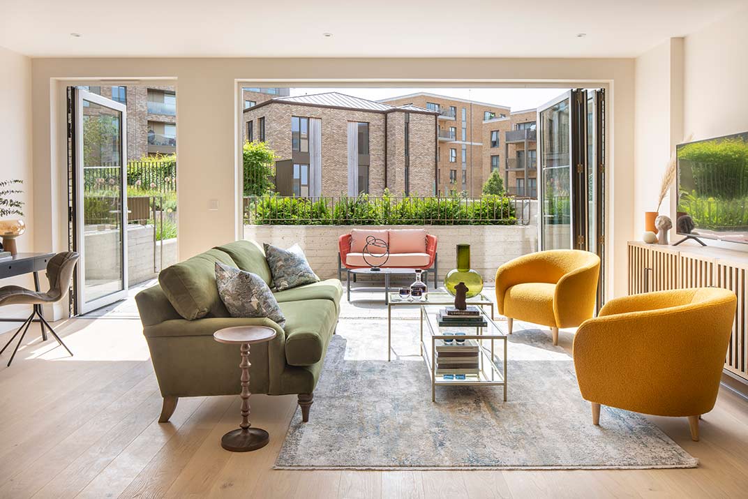 A modern living room with green sofa, two yellow armchairs, a glass coffee table, and large glass doors opening to a patio with outdoor seating and view of block of flats.