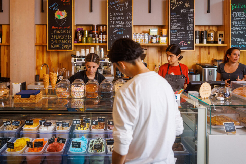 A person stands in front of a counter with various ice cream tubs, jars of toppings, and three staff members working behind the counter in a brightly lit ice cream parlour.