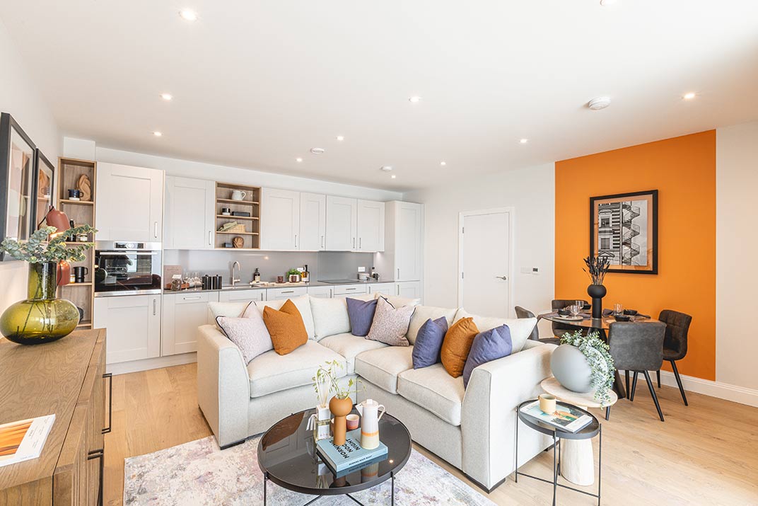 Modern open-plan living room and kitchen with white cupboards, a beige sofa with colourful cushions, round tables, and an orange feature wall behind a small dining area.