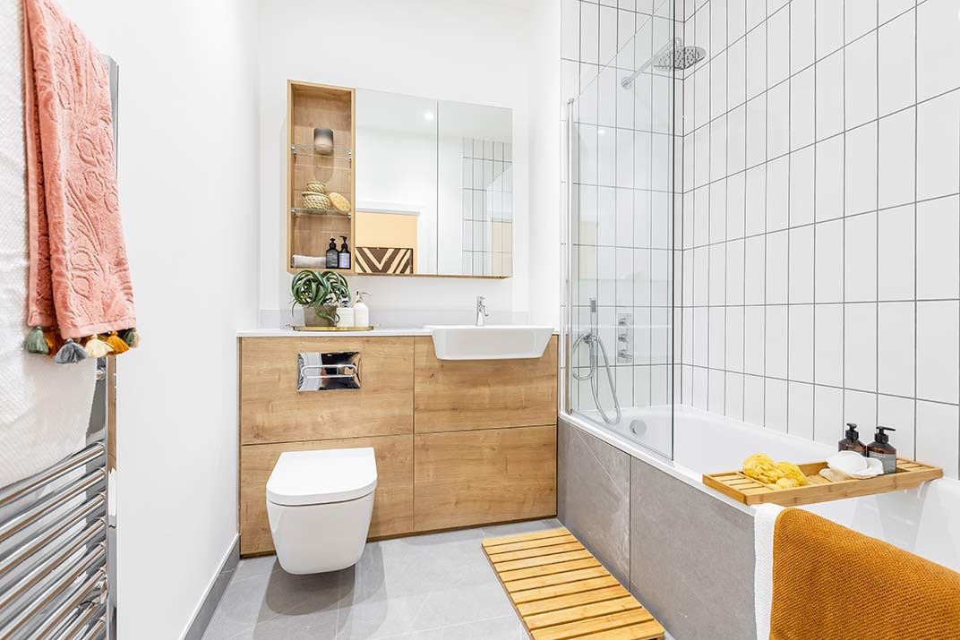Modern bathroom with wood-accented cupboards, white basin, wall-mounted toilet, bath with shower, towel rails, and decorative items on shelves.