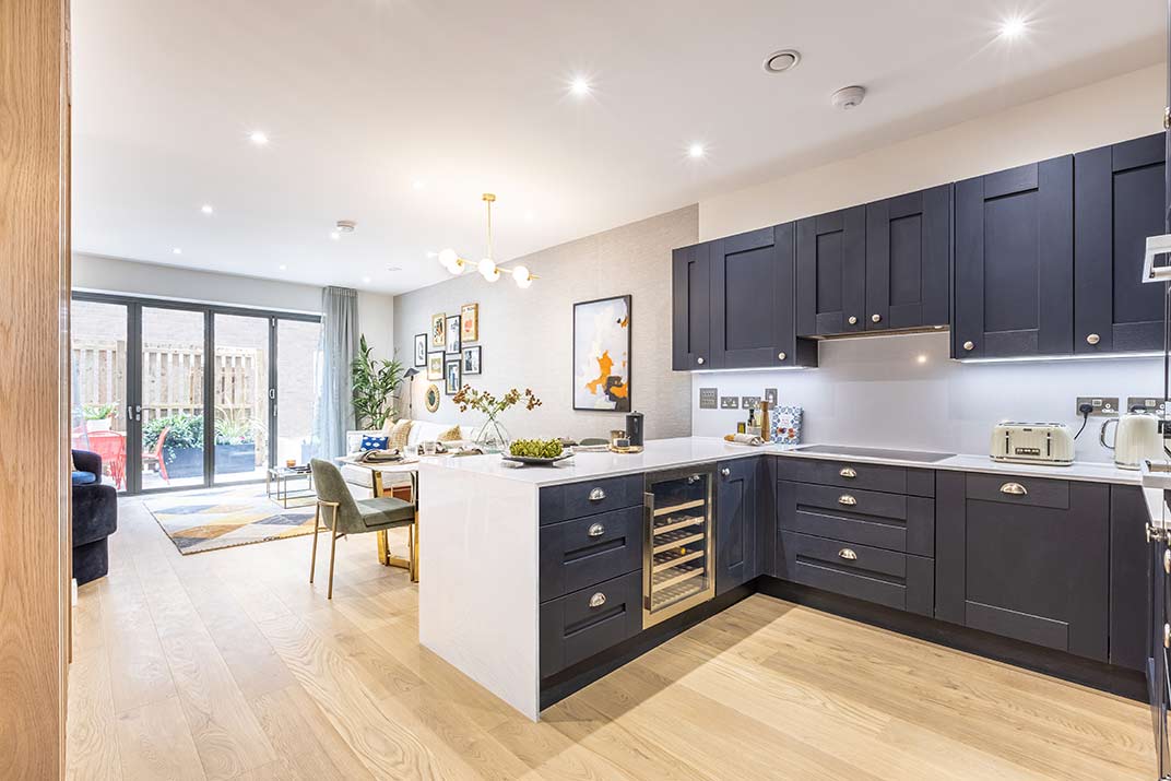 Modern open-plan kitchen and dining area with dark cupboards, built-in wine cooler, light wooden floors, and large windows opening to a patio.