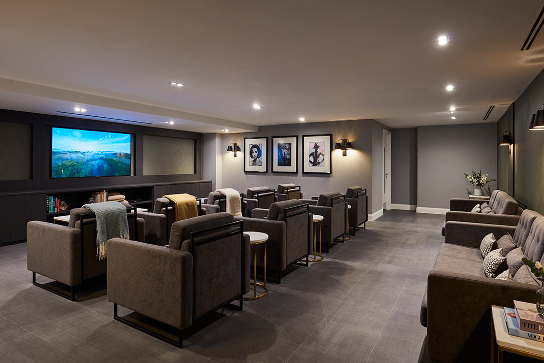 A modern home theatre room with rows of grey armchairs facing a large screen, side tables, wall art, and ambient lighting.