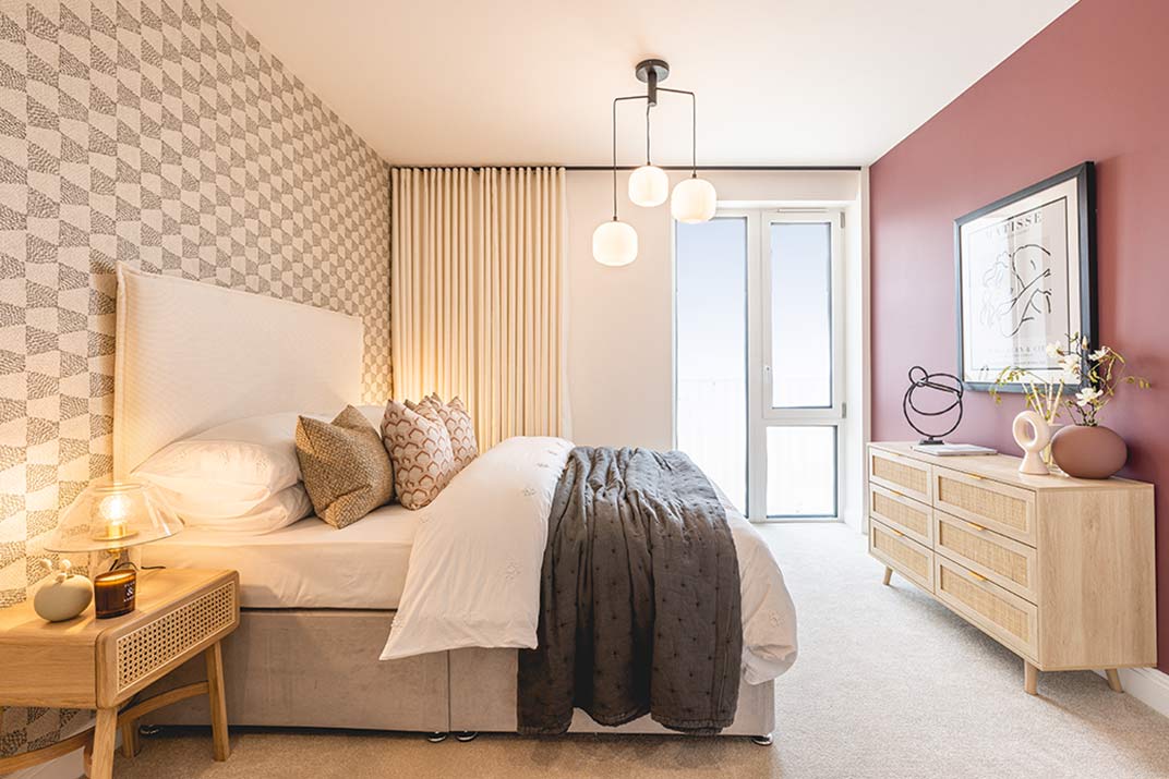 Modern bedroom with a double bed, geometric feature wall, pendant light fitting, wooden chest of drawers, and large window with sheer curtains.