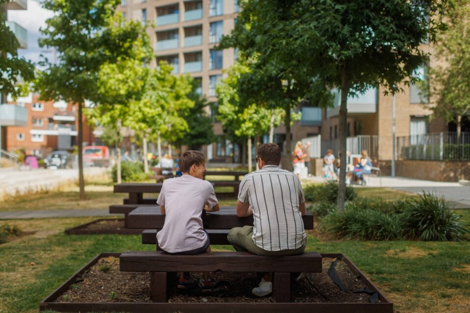 Two people sit on a wooden bench in an outdoor urban park area, facing away from the camera, with trees and blocks of flats in the background.
